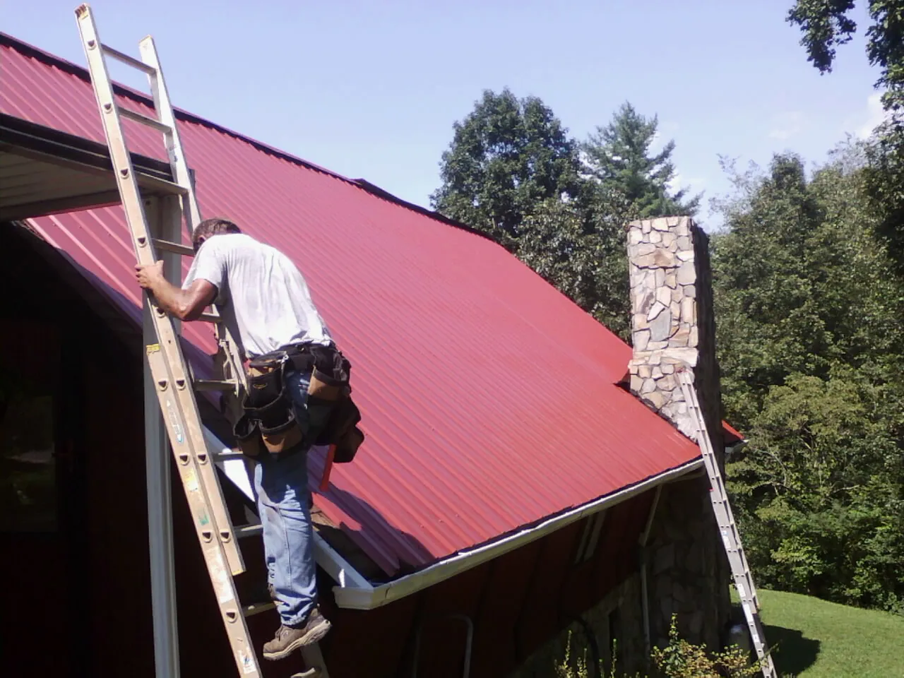 Roofer on a ladder beside a red metal roof and stone chimney — Soffit & Fascia Repair by Roofing Brothers in Beale Afb